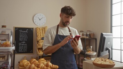 Young man in bakery shop celebrating with closed fist while using smartphone among display counter with pastries and bread shelves in background