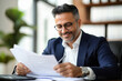 © Shivani - smiling businessman sitting at his desk in his office and reading a paper document