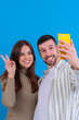 © alfons - Happy couple taking a selfie with a yellow smartphone against a blue background.