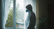 © GustavsMD - A healthcare professional in full protective gear, including a mask and suit, gazes out a window on a rainy day, symbolizing dedication and reflection