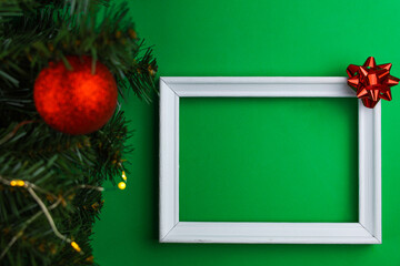 A white wooden frame lies on the side of the photo, with a red bow in the right corner and on the left of the image you can see the branches of a Christmas tree with a red shiny ball and a yellow