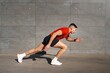 © Studio Marmellata - A man wearing a red shirt and black shorts sprints forward against a gray concrete wall under a clear blue sky with sunlight casting shadows