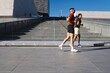 © Studio Marmellata - A man in a red shirt and a man in a black shirt jog side by side on a modern urban pathway with sleek architectural structures in the background under a clear blue sky
