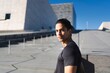 © Studio Marmellata - A man in a black shirt stands confidently outdoors with a modern architectural backdrop under a bright blue sky, looking directly at the camera
