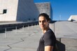© Studio Marmellata - A man in a black shirt stands confidently outdoors with a modern architectural backdrop under a bright blue sky, looking directly at the camera