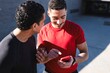 © Studio Marmellata - Two men in casual athletic wear share a moment of laughter as they look at a red smartphone together, standing outside on a sunny day with a concrete background
