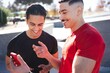 © Studio Marmellata - Two men in casual activewear laugh together while holding a smartphone, standing outdoors under the bright sun with trees and a modern background