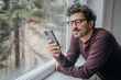 © Akbari - Young handsome middle eastern man scrolling on mobile phone in the morning standing near window.