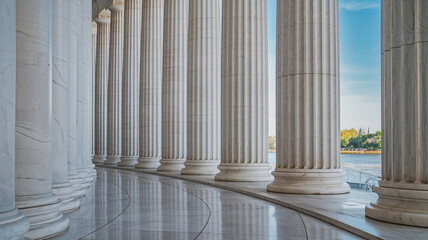 Tall classical white marble columns standing in a row with polished surfaces, marble ground with curved patterns leading to a serene view of water, distant trees, and faintly visible buildings 