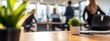 © David Giraud - Table en bois avec ordinateurs portables et plante verte sur un arrière-plan flou de gens travaillant dans un bureau d'entreprise.