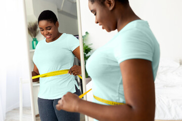 Wall Mural - Plus size millennial black woman measuring waist with tape, standing in front of mirror, satisfied with results of slimming diet at home. Young African American lady achieving success in weight loss