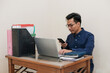 © Nurma - Smiling Southeast Asian Man Working While Sitting In Front Of Laptop and holding handphone In Home Office. Workplace Of Professional Worker, Freelancer or Student.
