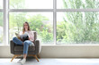 © Pixel-Shot - Young woman reading magazine while sitting in armchair near large window in room