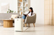 © Pixel-Shot - Modern air purifier on floor against woman reading book in armchair at home