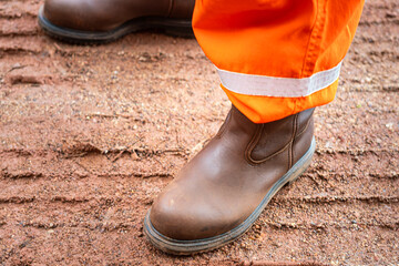 Wall Mural - A construction worker is wearing safety shoe and orange coverall uniform. He is standing on dirt ground at construction working site. Ready to work at unsafe workplace concept.