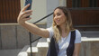 © Krakenimages.com - A young attractive blonde woman taking a selfie on an urban city street, smiling and dressed stylishly in casual wear with earrings and a vest under sunny outdoor conditions.