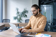 © Liubomir - A professional male employee concentrates on his work using a laptop in a bright office setting, exuding productivity and focus. The workspace includes modern furniture and a calm ambiance
