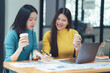 © Kritdanai - Teamwork Makes the Dream Work: Two smiling Asian businesswomen collaborate on a project, enjoying coffee break in a modern office setting.