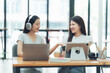 © Kritdanai - Sharing a Laugh: Two young women enjoy a lighthearted moment while working in a modern office setting, showcasing a sense of camaraderie and collaboration.