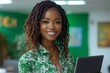 © serdon - A joyful African-American woman employee, standing in a contemporary office space with a laptop, looks at the camera with a light, friendly smile. She is a cheerful black businesswoman wearing a