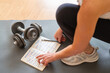 © Nanci - Female Athlete Organizing Fitness Plan On Notepad While Sitting On Yoga Mat With Dumbbells, Creating Healthy Daily And Weekly Exercise Routines, Promoting Active Lifestyle Goals, Morning Motivation