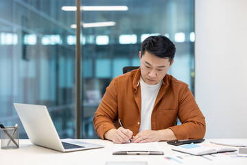 Poster - Asian man in a professional office environment writing notes while working with documents and a laptop, showcasing dedication and focus. Concept of business routine, office work, and productivity.