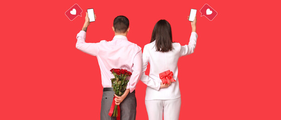 Wall Mural - Young couple with mobile phones, bouquet of roses and gift box on red background. Online dating