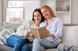 © Pixel-Shot - Pretty young woman and her mother with book sitting on sofa at home