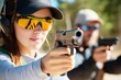 © LifeMedia - A woman wearing safety glasses and a cap aims a handgun at a target on an outdoor shooting range, demonstrating concentration and precision in firearm handling.
