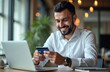 © miss irine - Arabian businessman smiles happily using mobile phone, credit card for online shopping at modern office desk. Focused on phone, card in hand. Businessman wearing white shirt. Sits in well lit office.