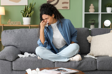 Wall Mural - Young African-American woman sneezing in handkerchief and sitting on sofa at home. Allergy concept