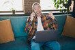 © deagreez - Young man celebrating success at home while talking on the phone and using a laptop on a cozy couch in a stylish living room