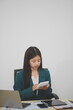 © Sirikarn Rinruesee - Asian businesswoman in red suit calculating company sales with calculator, laptop, and tablet on table Interior of businesswoman office at the modern workstation.