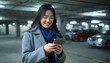 © Flash - Asian woman in gray coat, blue scarf, smiling, holding smartphone, in dimly lit parking garage with parked cars