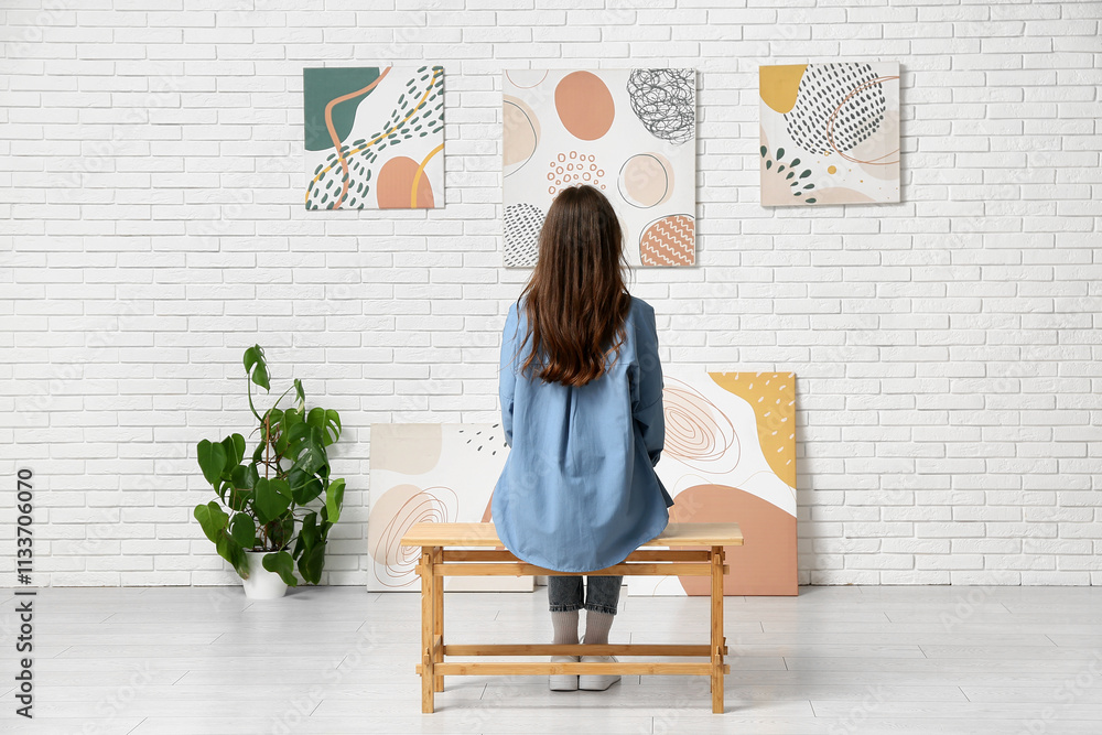 Young woman on bench looking at paintings in art gallery, back view