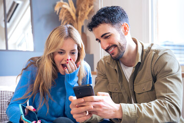 a young couple seated indoors sharing a surprising and joyful moment while looking at a smartphone. 