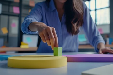 Leinwandbilder - Close-up of a woman's hand placing a green cube on a yellow circle, part of a colorful geometric arrangement on a desk.