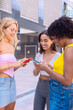 © Raquel - Three cheerful young women gathers around a red smartphone, reacting with excitement. They are dressed casually, standing outdoors in a modern urban environment, enjoying a fun moment together.