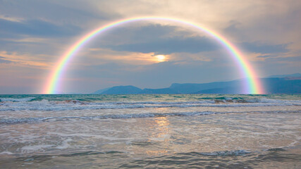 Poster - A rainbow over a beach with waves crashing against the shore, under a dramatic sky.