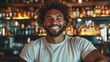 © familymedia - A bartender with curly hair exuberantly smiles in a cozy bar with shelves of bottles, creating a warm and inviting atmosphere for patrons and visitors alike.