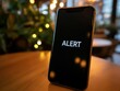 © Andrew - Urgent Smartphone Alert:  A dramatic close-up shot of a smartphone displaying a stark 'ALERT' message on its screen, resting on a wooden table in a softly lit cafe setting.