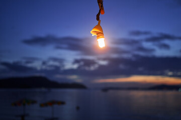 Sticker - A single light bulb hanging against a twilight sky with clouds and distant mountains.