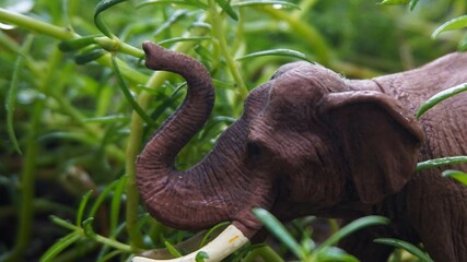 elephant toy, cute elephant miniature,A detailed close-up of a realistic small elephant model surrounded by green foliage. This brown elephant model has a protruding trunk and partially visible tusks,