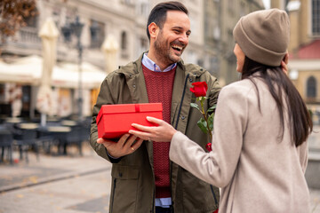 a mid adult caucasian male surprises his partner with a red rose on a city street. dressed casually,