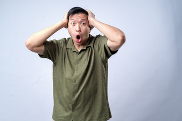 An Asian man in a casual green shirt holds his head with both hands and opens his mouth wide in shock or surprise, standing against a clean white background. The image conveys surprise