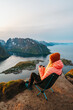 © EVERST - Outdoor travel hiking adventure vacations in Norway - woman relaxing in camping chair with thermos on Reinebringen mountain top enjoying aerial view of Reine village and fjord in Lofoten Islands