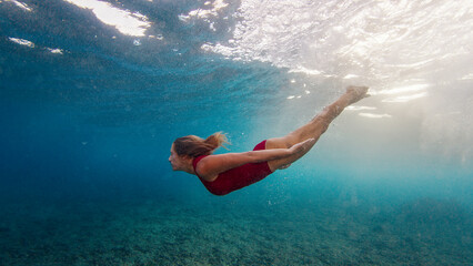 Wall Mural - Young slim woman in red suit dives under the breaking wave and swims confidently underwater in the harsh sea environment