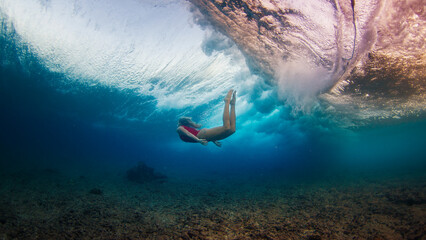 Wall Mural - Young slim woman in red suit dives under the breaking wave and swims confidently underwater in the harsh sea environment