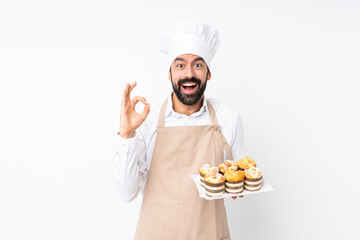 Wall Mural - Young man holding muffin cake over isolated white background surprised and showing ok sign