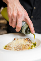 Wall Mural - A close-up of a chef's hand drizzling green oil over a plated dish topped with grated cheese. The focus is on the gourmet presentation and meticulous attention to detail in the kitchen.